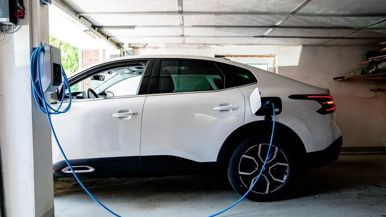 Vit elbil laddas i garage. En blå laddningskabel går från väggen till bilen. Tänk på brandrisk vid laddning av elbil i garage.
