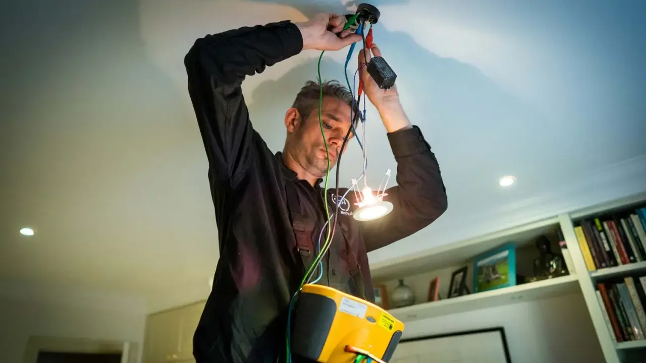 En auktoriserad elektriker kopplar in en ny taklampa. Han har en gul mätutrustning runt halsen.