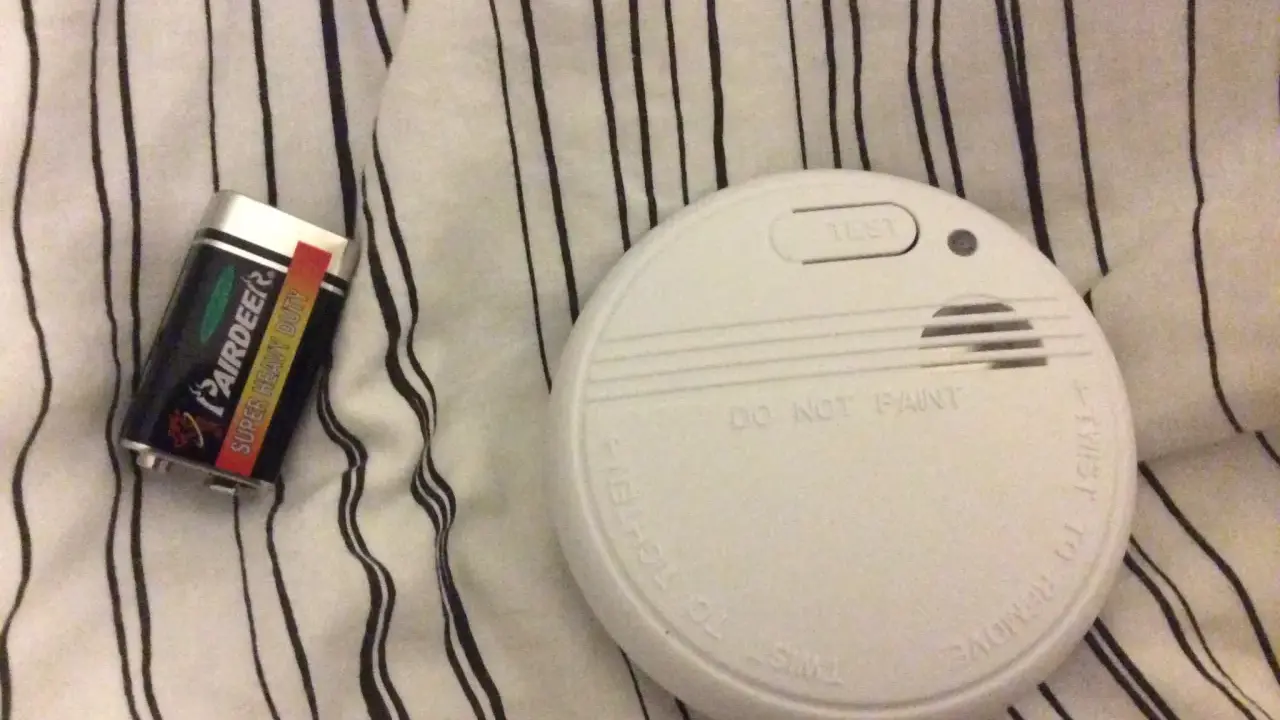Ett brandlarm och ett batteri ligger på en randig duk. Brandlarmet piper inte, men det är redo att varna vid fara.