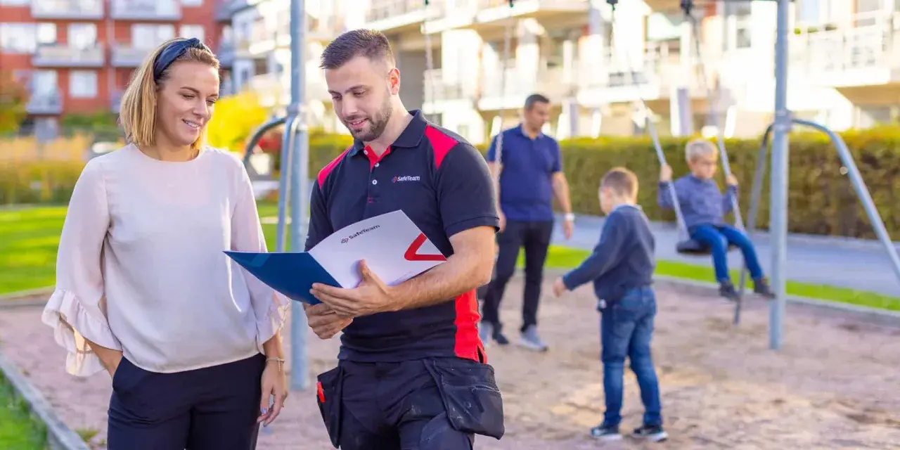 En man och en kvinna diskuterar dokument i en lekpark. Mannen, klädd i arbetskläder, visar upp information som kan relatera till boverkets byggregler.
