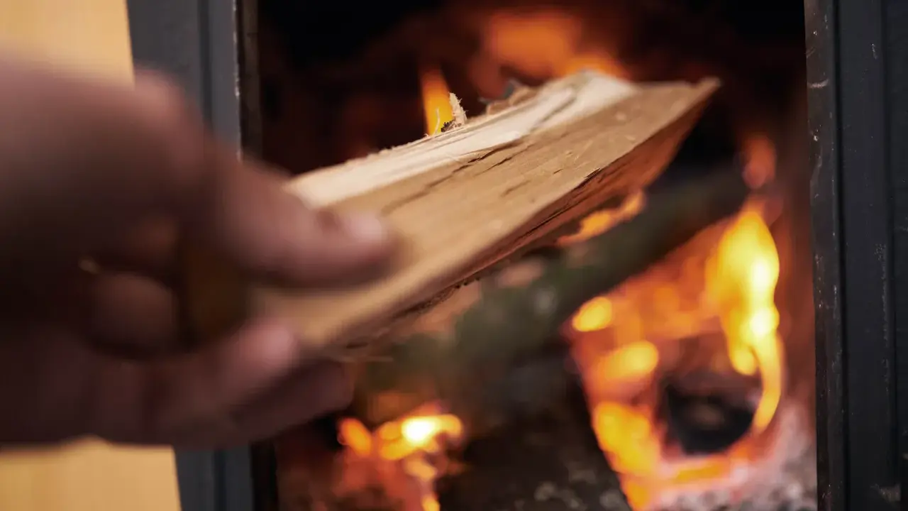En hand lägger ved i en kamin där elden flammande ryker in från.
