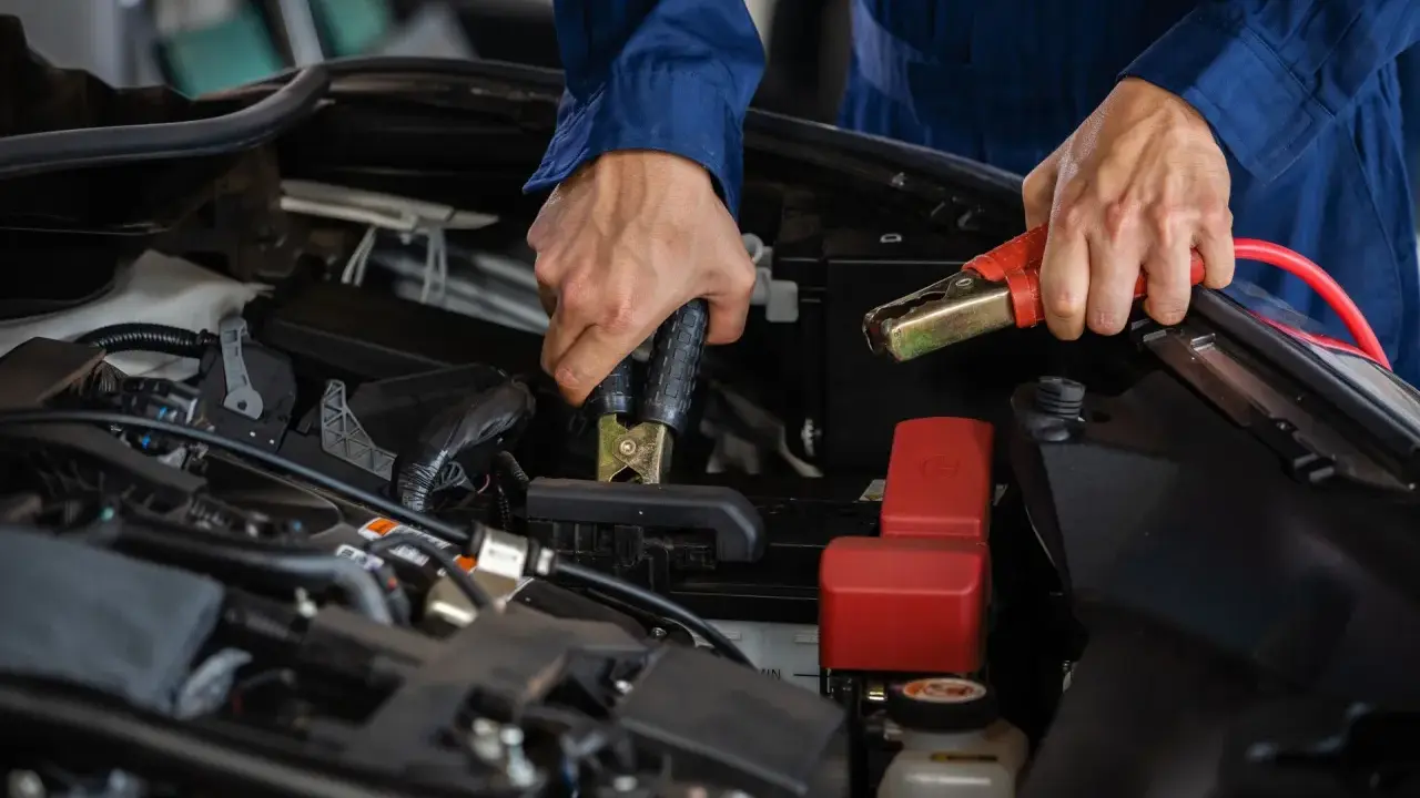 Person i blå overall kopplar startkablar för att ladda bilbatteri inomhus.