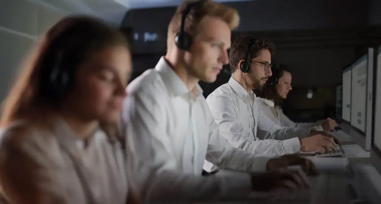 Personal som arbetar på en **Nokas larmcentral** i mörker, fokuserade på sina datorskärmar med headset på.