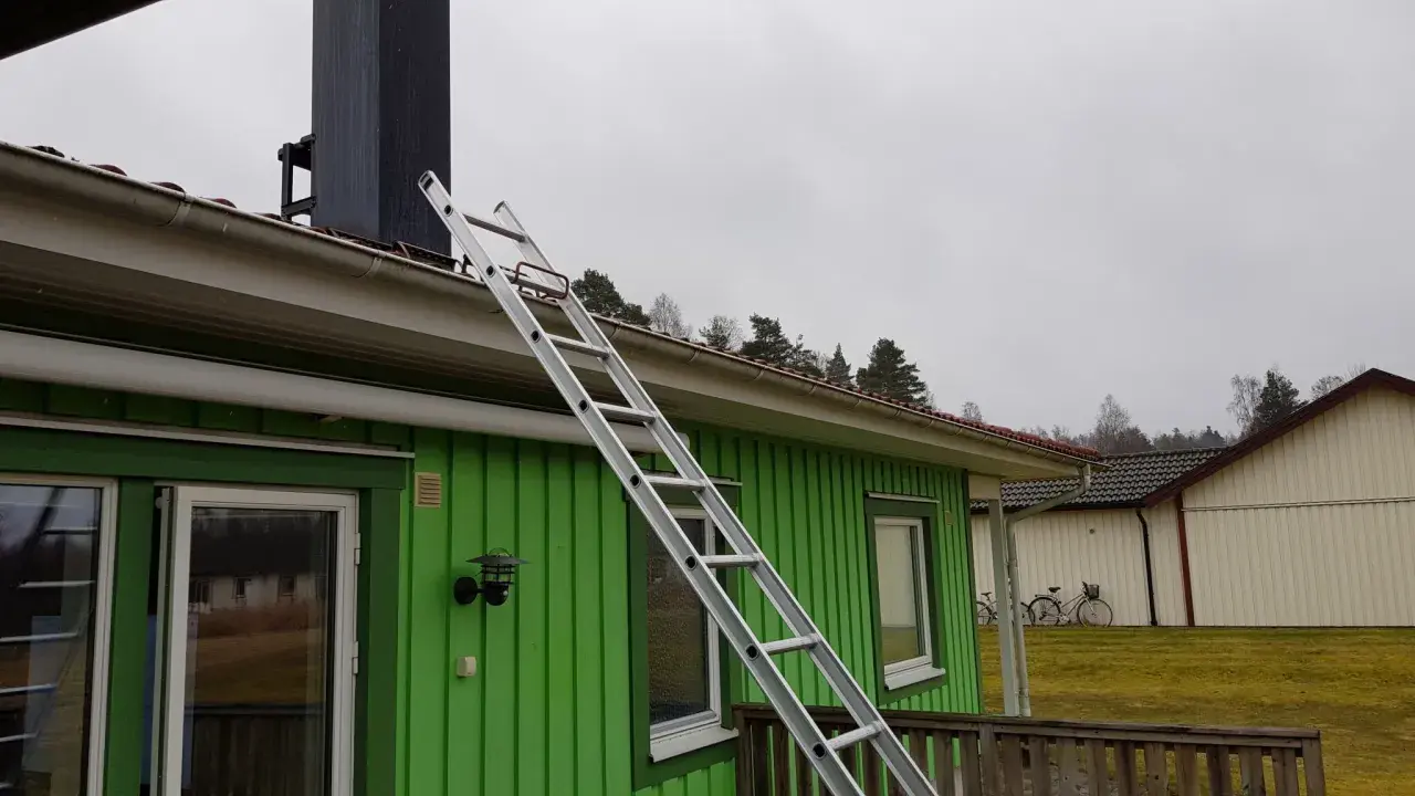 En aluminiumstege lutar mot taket på ett grönt hus, redo för arbete vid skorstenen. En krok till stege är synlig.