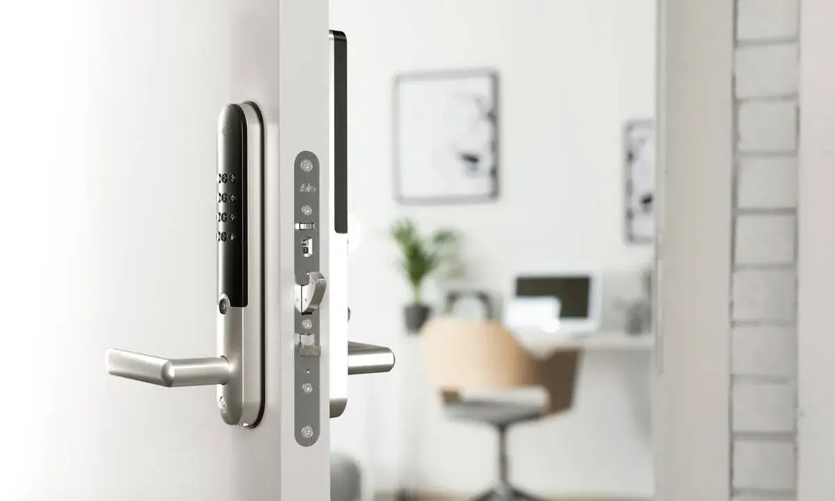 Ett modernt, silverfärgat assa elektroniskt lås med kodpanel och handtag på en vit dörr. I bakgrunden syns ett kontor.
