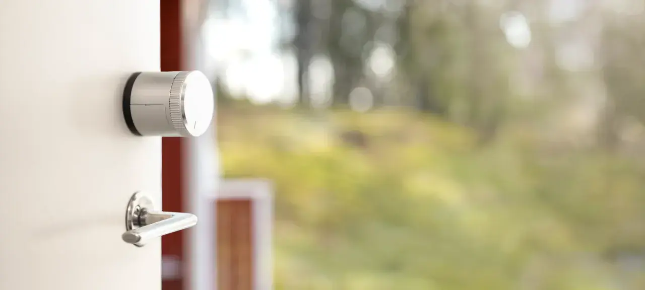En vit dörr med en silverfärgad Verisure smart låsmodul och ett dörrhandtag. Utanför syns en suddig grönskande trädgård.
