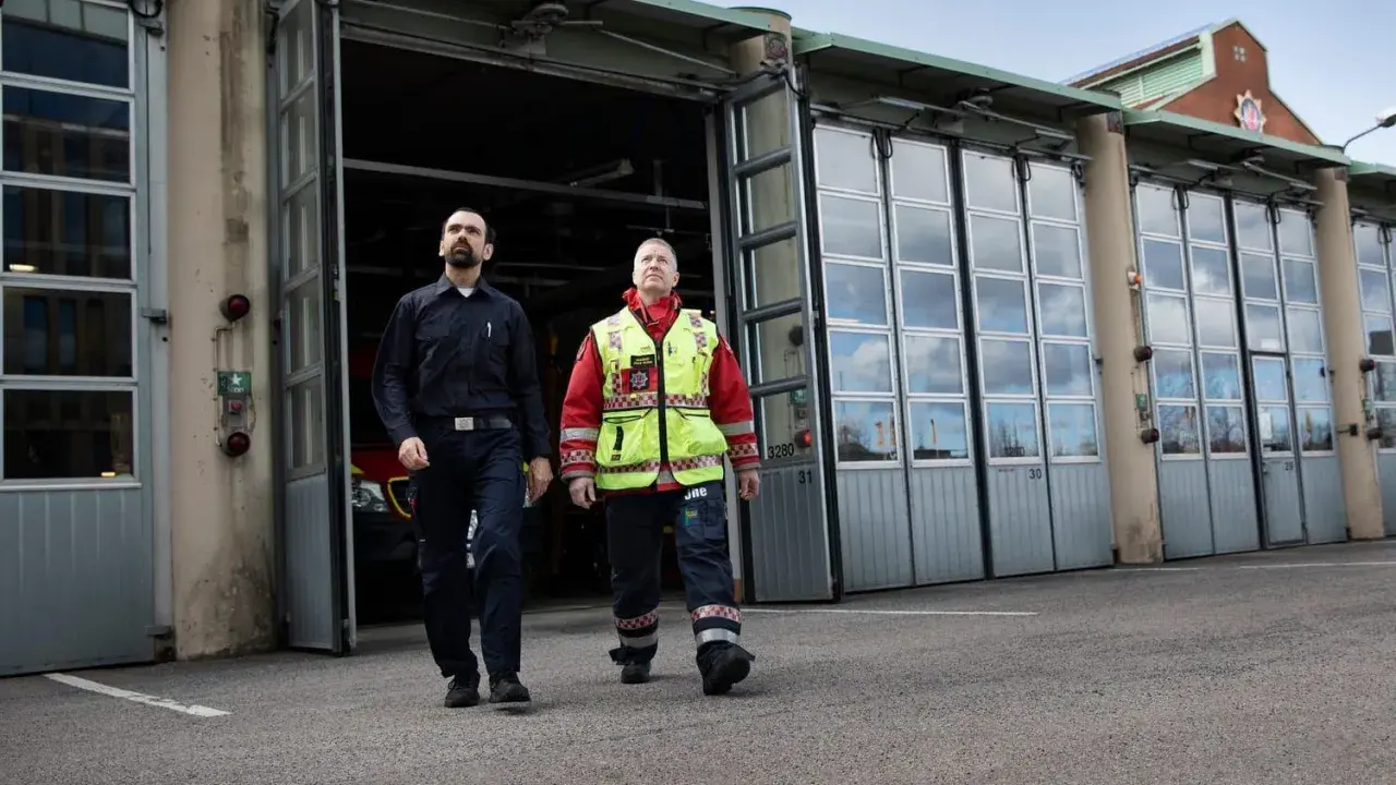 Två män går framför en brandstation. Den ena bär mörkblå uniform, den andra en gul reflexväst över röd jacka. De följer en stigarledning.