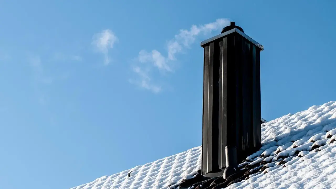 En svart skorsten av plåt ryker på ett snötäckt tak mot en klarblå himmel.