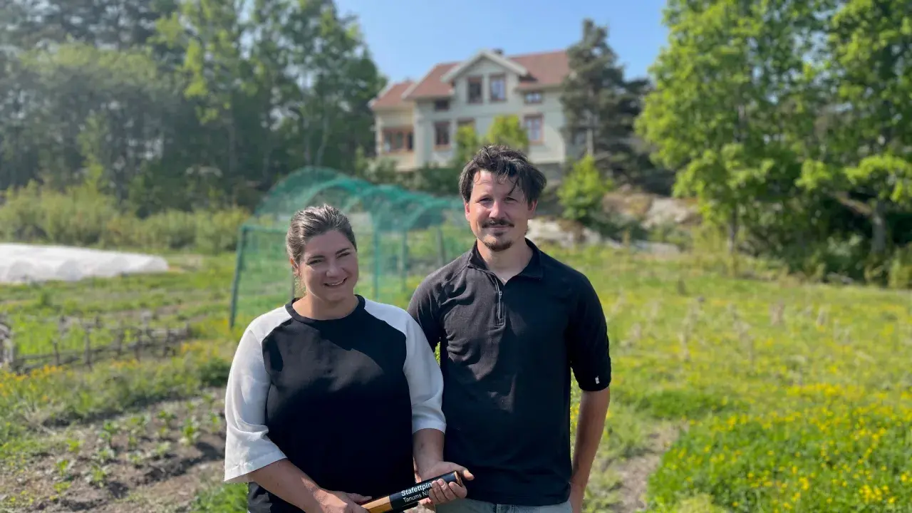 Två personer står på ett fält med ett hus i bakgrunden. De arbetar med lbk lantbruk och visar upp en produkt.
