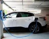 Vit elbil laddas i garage. En blå laddningskabel går från väggen till bilen. Tänk på brandrisk vid laddning av elbil i garage.