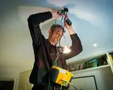 En auktoriserad elektriker kopplar in en ny taklampa. Han har en gul mätutrustning runt halsen.