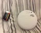 Ett brandlarm och ett batteri ligger på en randig duk. Brandlarmet piper inte, men det är redo att varna vid fara.