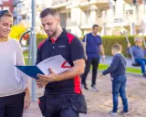 En man och en kvinna diskuterar dokument i en lekpark. Mannen, klädd i arbetskläder, visar upp information som kan relatera till boverkets byggregler.