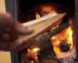 En hand lägger ved i en kamin där elden flammande ryker in från.