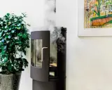 En modern svart stående kamin med glasdörr och synliga vedträn. Rök stiger från skorstenen, en grön växt står bredvid.