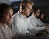 Personal som arbetar på en **Nokas larmcentral** i mörker, fokuserade på sina datorskärmar med headset på.