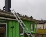 En aluminiumstege lutar mot taket på ett grönt hus, redo för arbete vid skorstenen. En krok till stege är synlig.