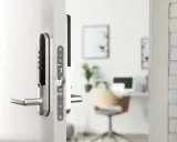 Ett modernt, silverfärgat assa elektroniskt lås med kodpanel och handtag på en vit dörr. I bakgrunden syns ett kontor.