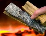 Hand håller vedbitar med bark, redo att elda med fuktig ved. Flammor syns i bakgrunden.