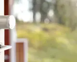 En vit dörr med en silverfärgad Verisure smart låsmodul och ett dörrhandtag. Utanför syns en suddig grönskande trädgård.