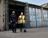 Två män går framför en brandstation. Den ena bär mörkblå uniform, den andra en gul reflexväst över röd jacka. De följer en stigarledning.