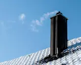 En svart skorsten av plåt ryker på ett snötäckt tak mot en klarblå himmel.