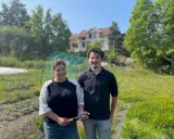 Två personer står på ett fält med ett hus i bakgrunden. De arbetar med lbk lantbruk och visar upp en produkt.
