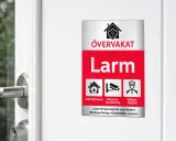 En larmskylt utan larm, men med symboler för inbrottslarm, kameraövervakning och väktaråtgärd, sitter på en vit dörr.