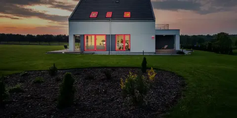 Modern villa brand med lysande röda fönsterramar mot en dramatisk solnedgång.