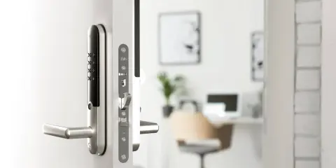 Ett modernt, silverfärgat assa elektroniskt lås med kodpanel och handtag på en vit dörr. I bakgrunden syns ett kontor.