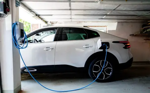 Vit elbil laddas i garage. En blå laddningskabel går från väggen till bilen. Tänk på brandrisk vid laddning av elbil i garage.