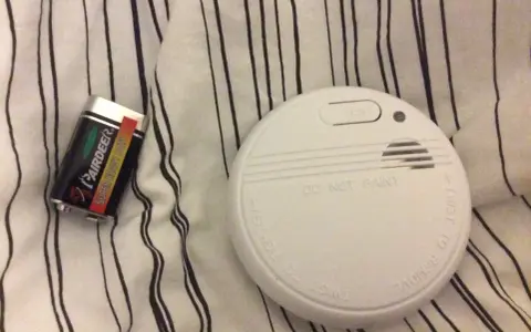 Ett brandlarm och ett batteri ligger på en randig duk. Brandlarmet piper inte, men det är redo att varna vid fara.