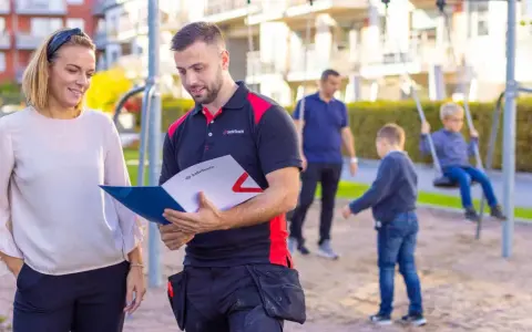 En man och en kvinna diskuterar dokument i en lekpark. Mannen, klädd i arbetskläder, visar upp information som kan relatera till boverkets byggregler.