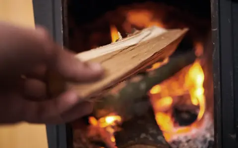 En hand lägger ved i en kamin där elden flammande ryker in från.