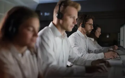 Personal som arbetar på en **Nokas larmcentral** i mörker, fokuserade på sina datorskärmar med headset på.