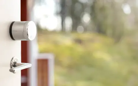 En vit dörr med en silverfärgad Verisure smart låsmodul och ett dörrhandtag. Utanför syns en suddig grönskande trädgård.