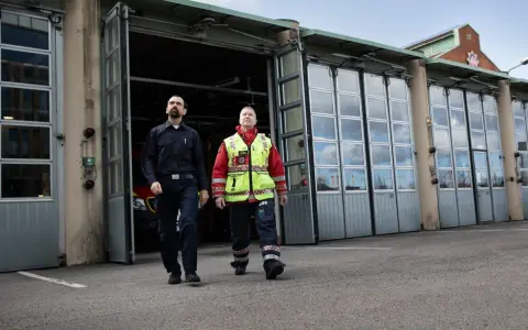 Två män går framför en brandstation. Den ena bär mörkblå uniform, den andra en gul reflexväst över röd jacka. De följer en stigarledning.