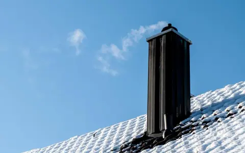 En svart skorsten av plåt ryker på ett snötäckt tak mot en klarblå himmel.