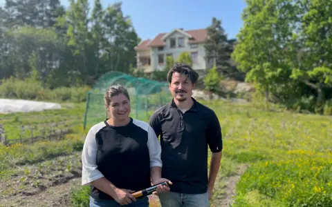 Två personer står på ett fält med ett hus i bakgrunden. De arbetar med lbk lantbruk och visar upp en produkt.