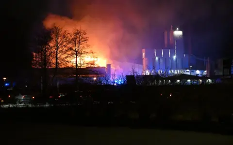 En fabrik brinner med orange och lila rök. Kanske en påminnelse om vikten av att följa kurs Boverkets brandregler för att undvika sådana katastrofer.