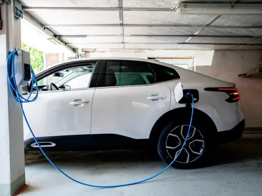 Vit elbil laddas i garage. En blå laddningskabel går från väggen till bilen. Tänk på brandrisk vid laddning av elbil i garage.