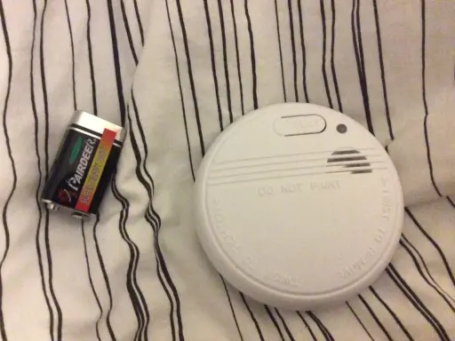 Ett brandlarm och ett batteri ligger på en randig duk. Brandlarmet piper inte, men det är redo att varna vid fara.