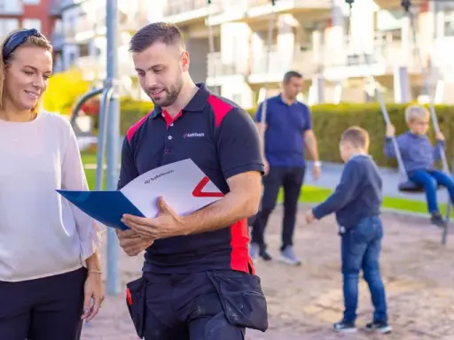 En man och en kvinna diskuterar dokument i en lekpark. Mannen, klädd i arbetskläder, visar upp information som kan relatera till boverkets byggregler.