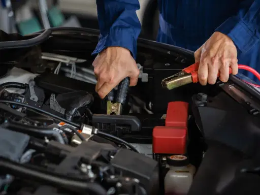 Person i blå overall kopplar startkablar för att ladda bilbatteri inomhus.