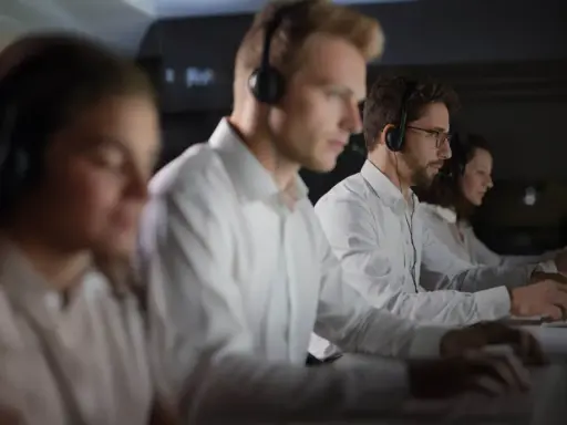 Personal som arbetar på en **Nokas larmcentral** i mörker, fokuserade på sina datorskärmar med headset på.