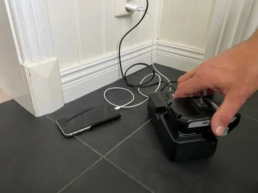 En hand placerar ett litiumbatteri på en laddare. En mobiltelefon laddas bredvid.