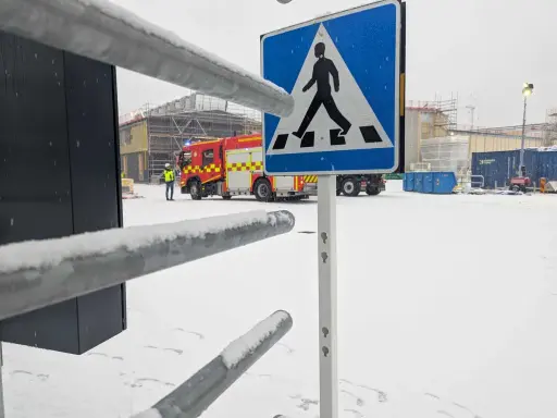 Snöig arbetsplats med brandbil och en person i varselväst. En övergångsställe-skylt står framför. Kanske en leveransbesiktning brandlarm pågår.