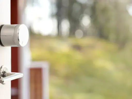 En vit dörr med en silverfärgad Verisure smart låsmodul och ett dörrhandtag. Utanför syns en suddig grönskande trädgård.