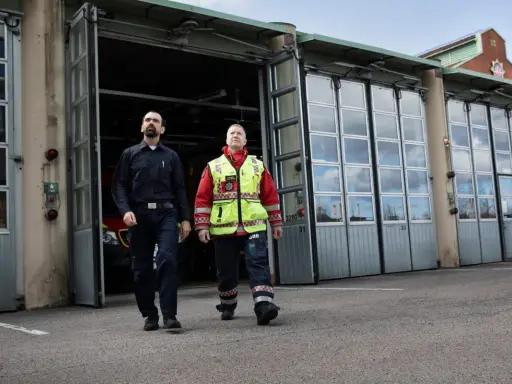 Två män går framför en brandstation. Den ena bär mörkblå uniform, den andra en gul reflexväst över röd jacka. De följer en stigarledning.