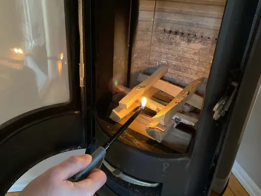 En hand tänder eld i kaminen med en lång tändare. Snart sprider sig värmen i rummet.